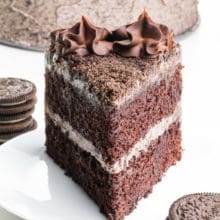 A slice of chocolate cake sits on a plate with chocolate sandwich cookies stacked in front and behind it. The rest of the cake is behind it too.