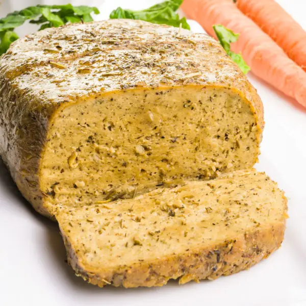 A vegan turkey loaf has a slice cut out of it. There are carrots in the background.