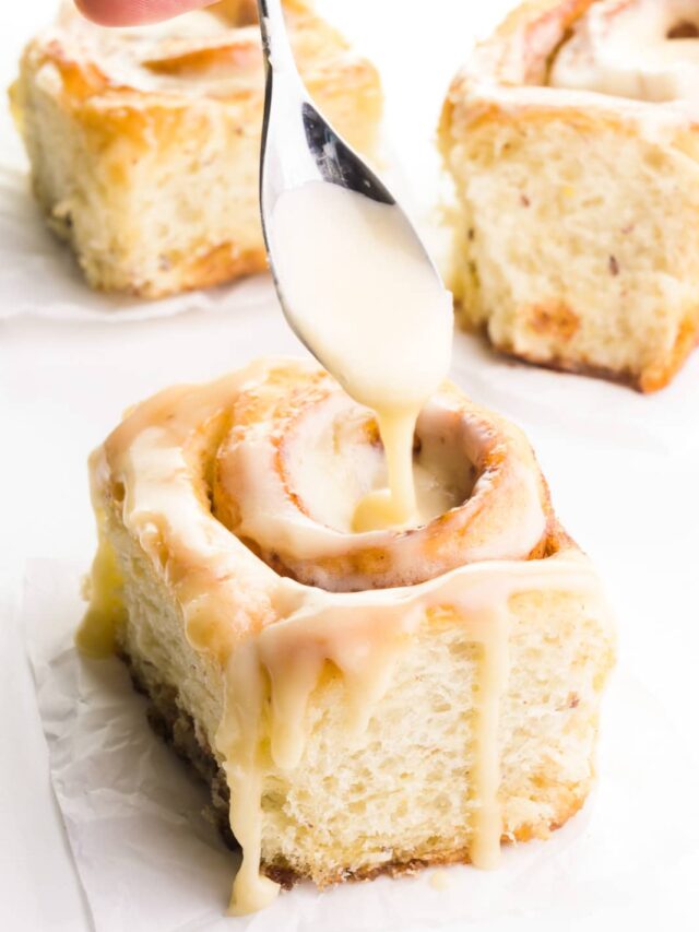 Three vegan cinnamon rolls are on a counter. A hand holds a spoon drizzling frosting over the top of one of them.