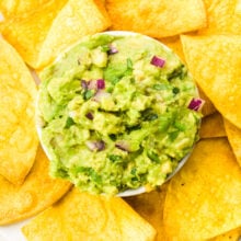 Looking down on a bowl of guacamole surrounded by air fried tortilla chips. There are cherry tomatoes and fresh limes nearby.