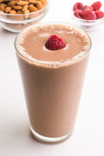A tall glass holds chocolate almond milk smoothie with a raspberry on top. There is a bowl of of almonds in the background sitting beside a bowl of fresh raspberries.