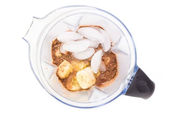 Looking down on a blender jar with ice cubes, banana chunks, and chocolate protein powder,