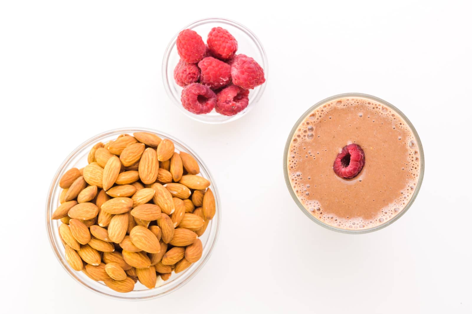 Looking down on a chocolate smoothie with a raspberry on top. There is a bowl of raspberries and a bowl of almonds next to it.