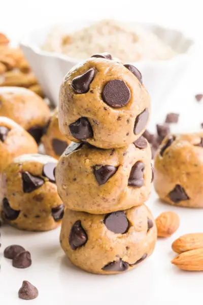 A stack of three almond butter balls are studded with chocolate chips. There are more balls in the background, along with almonds, chocolate chips, and a bowl of almond flour.
