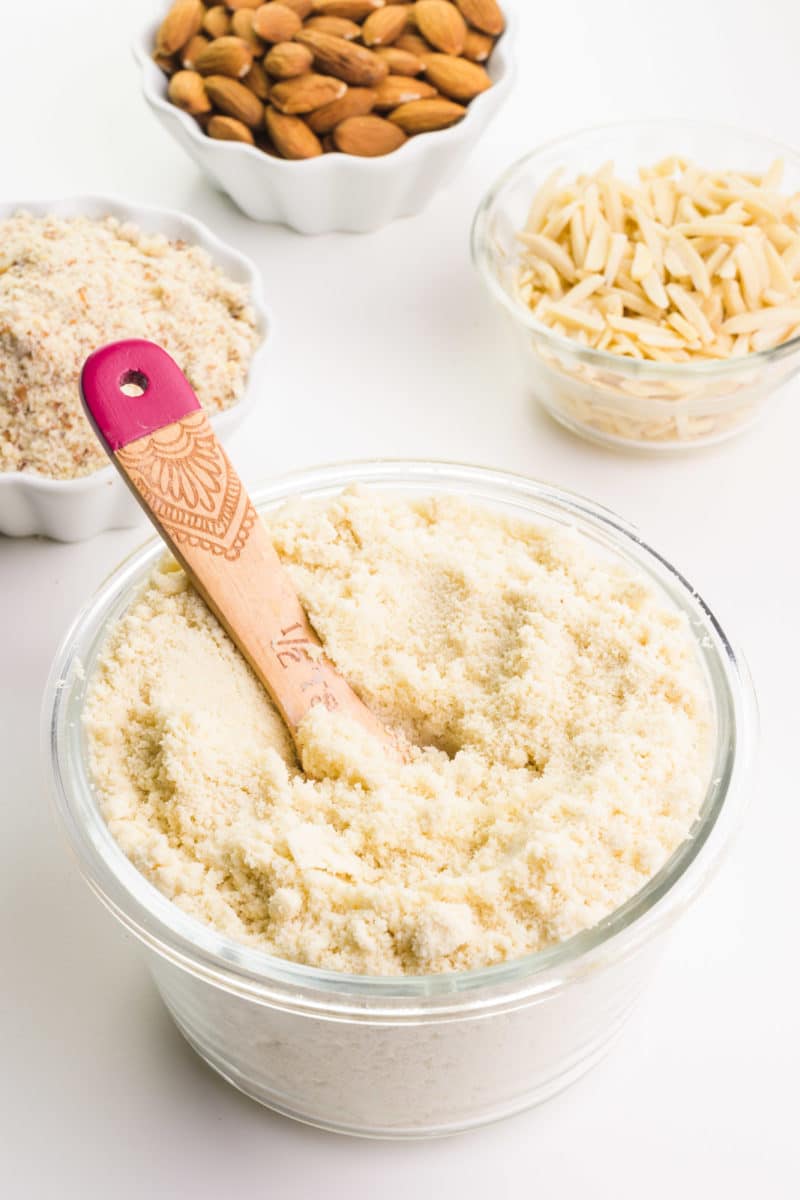 A wooden measuring spoon is in a bowl of almond flour. There are bowls in the background holding almond meal, raw almonds, and almond slivers.