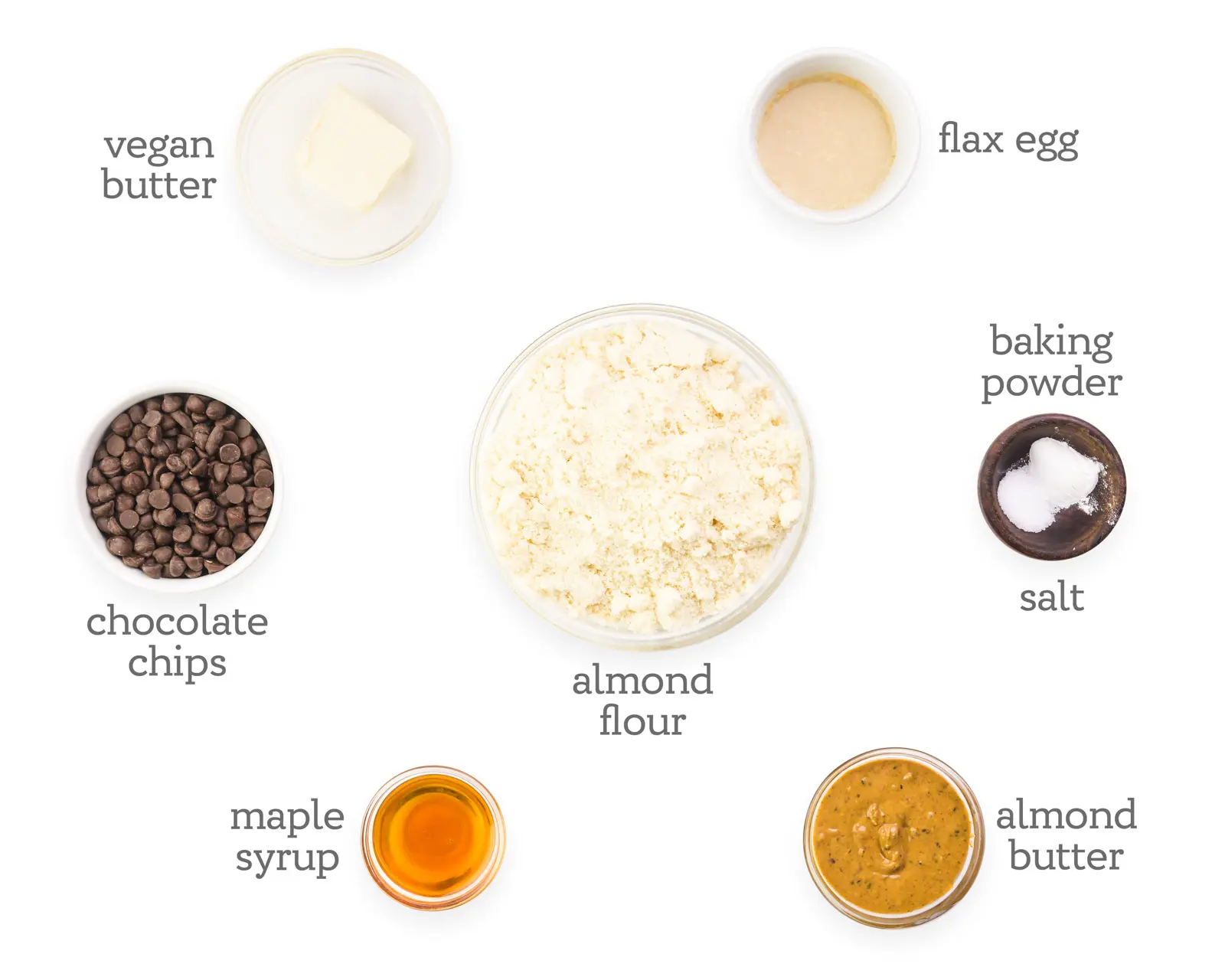 Ingredients in bowls are laid out on a white table. The ingredients next to the ingredients read, flax egg, baking powder, salt, almond butter, almond flour, maple syrup, chocolate chips, and vegan butter.