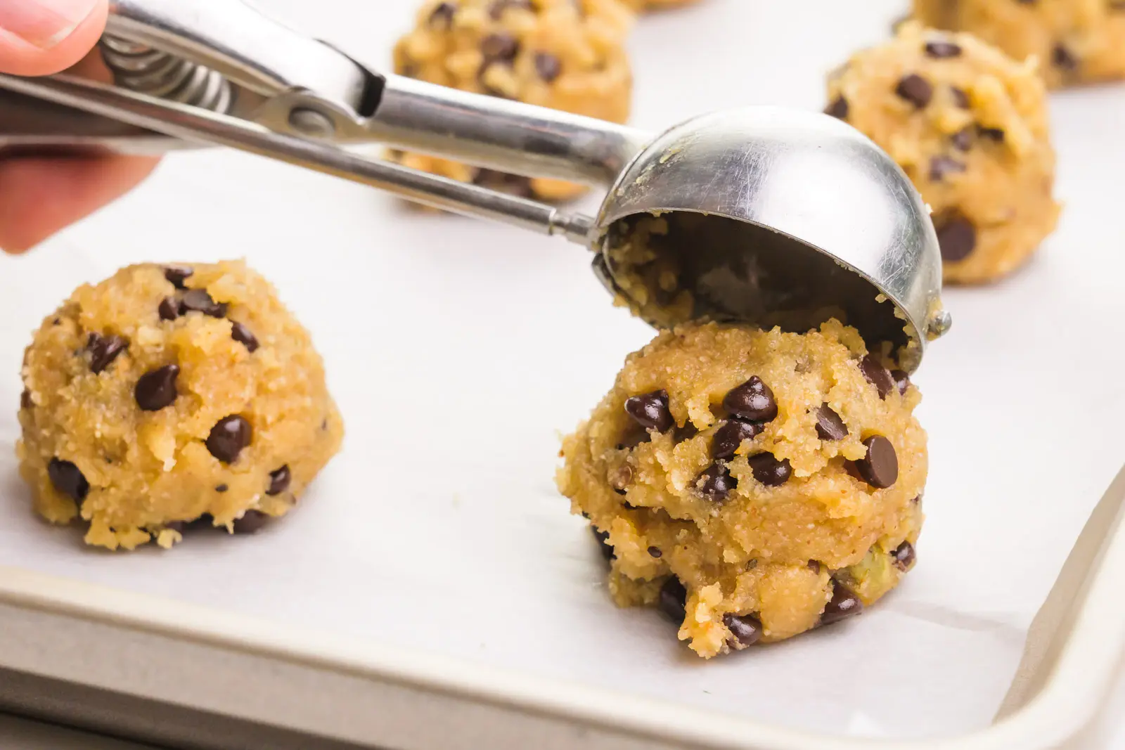 A hand holds a cookie scoop, scooping mounds of cookie dough batter on a cookie sheet lined with parchment paper.