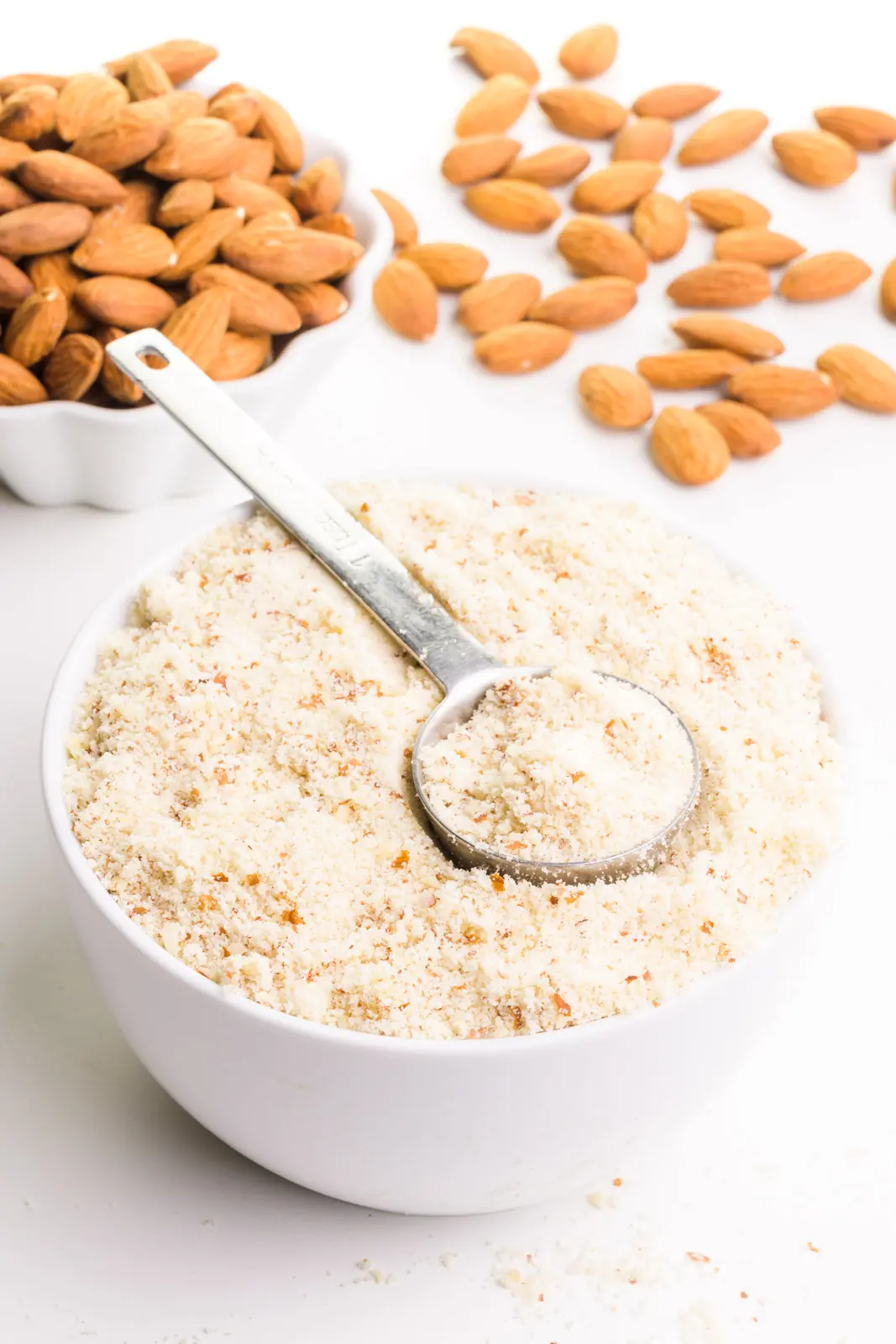 A metal measuring spoon sits in a bowl full of almond meal flour. There's a bowl of almonds in the background with more almonds on the table beside it.