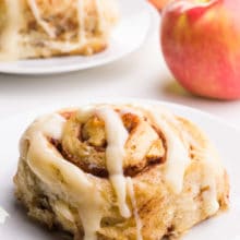 An apple cinnamon roll sits on a plate in front of another one on a plate in the background. There are two apples in the background.