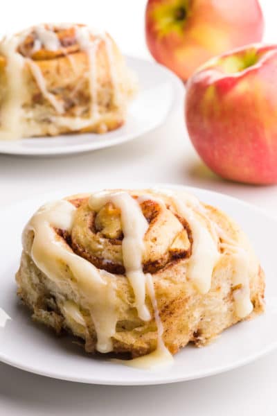 An apple cinnamon roll sits on a plate in front of another one on a plate in the background. There are two apples in the background.