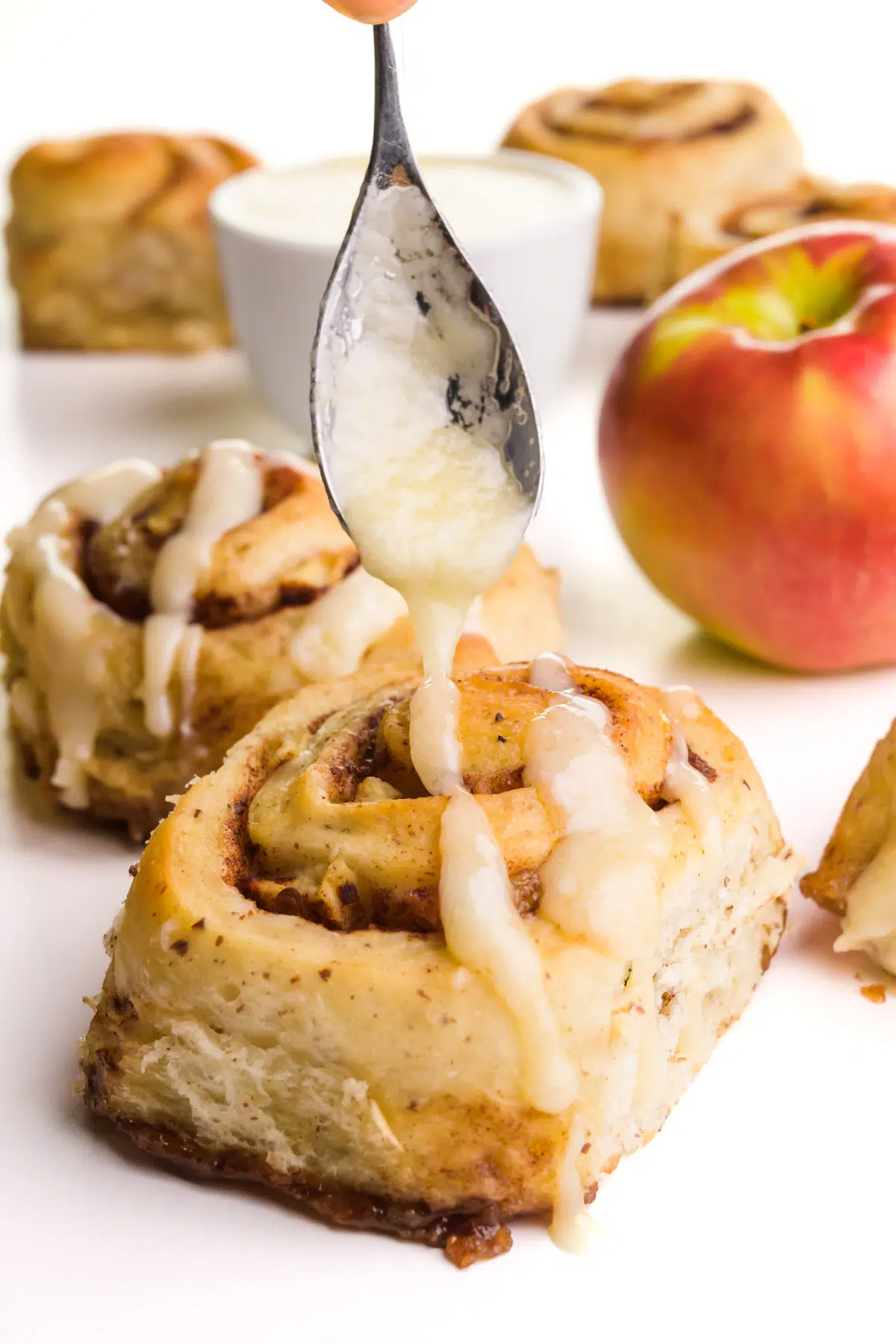 A spoon hovers over cinnamon rolls, drizzling icing on top. There is an apple and a bowl of icing in the background.