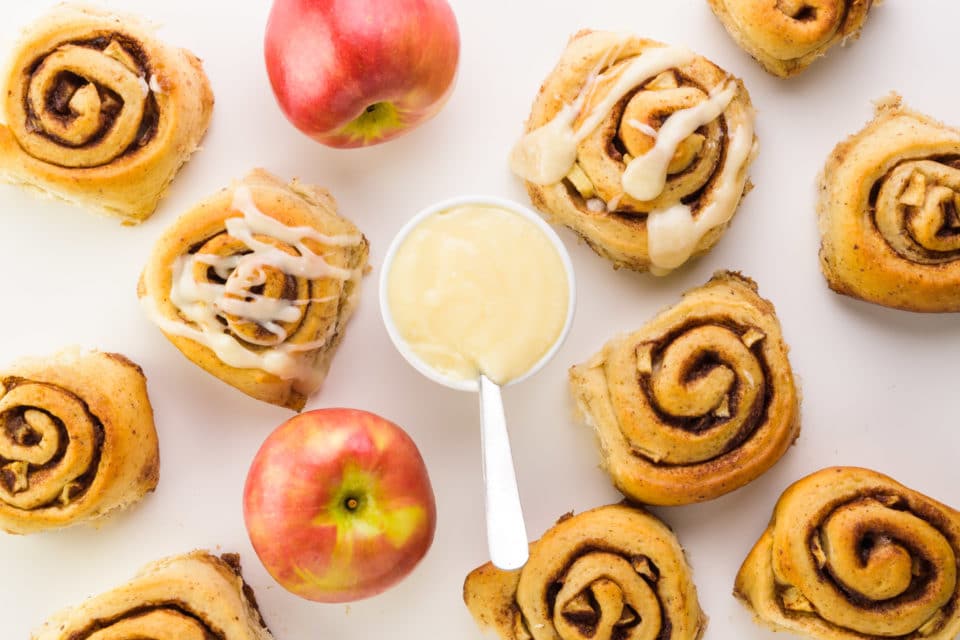 Looking down on several apple pie cinnamon rolls with a bowl of icing and some apples in the mix.