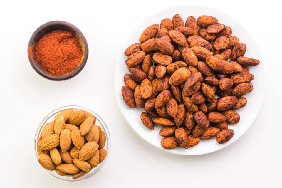 Looking down on a plate full of baked almonds. There's a bowl of raw almonds and a bowl of paprika next to it.