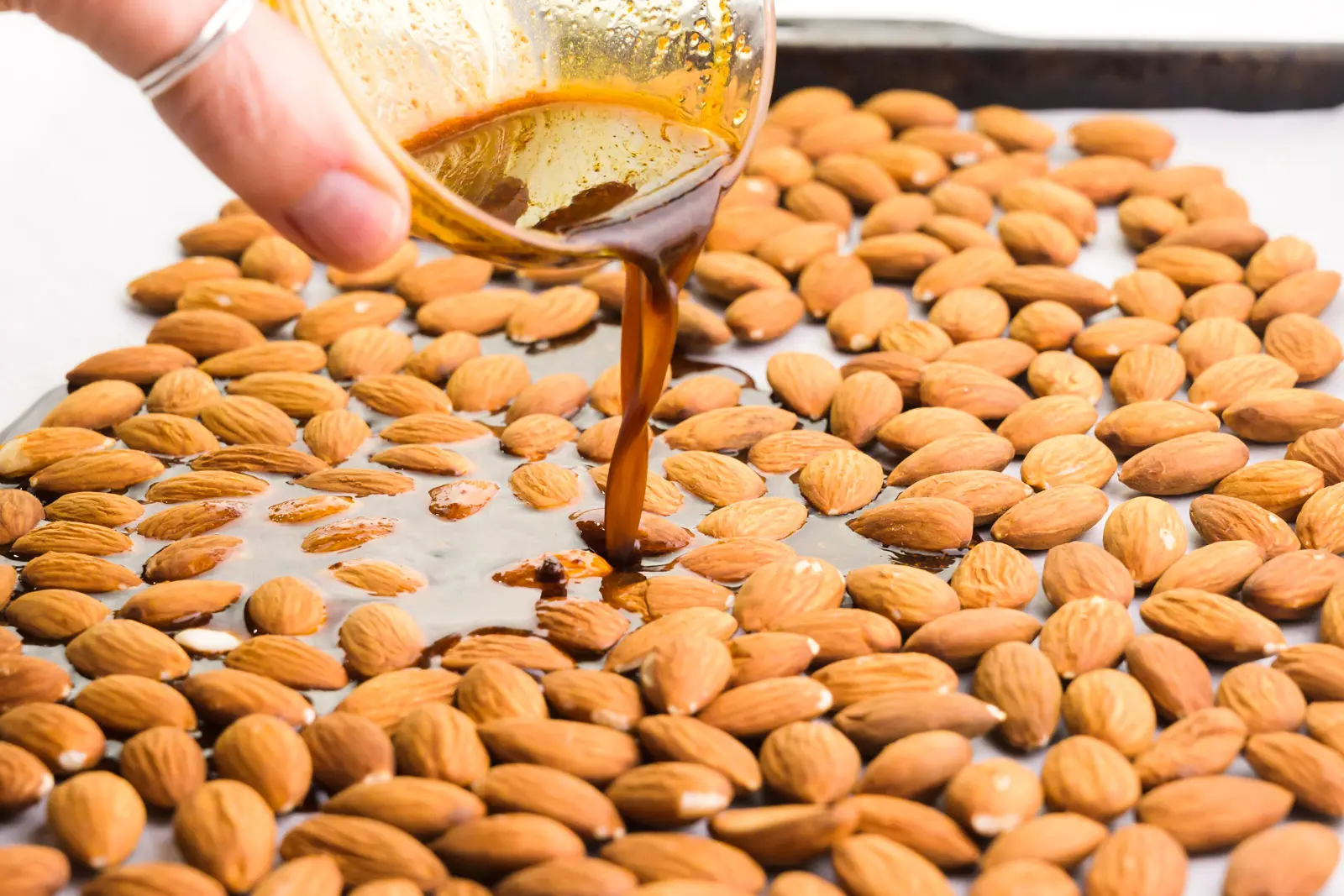 A hand holds a bowl and pouring sauce over a pan full of almonds.