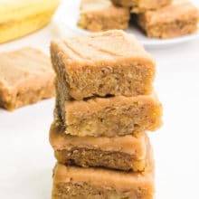 A closeup of a stack of banana blondies with peanut butter frosting. There are more slices and bananas in the background.