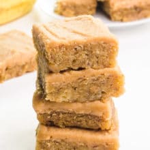 A stack of banana blondies with peanut butter frosting sits in front of more slices and bananas in the background.