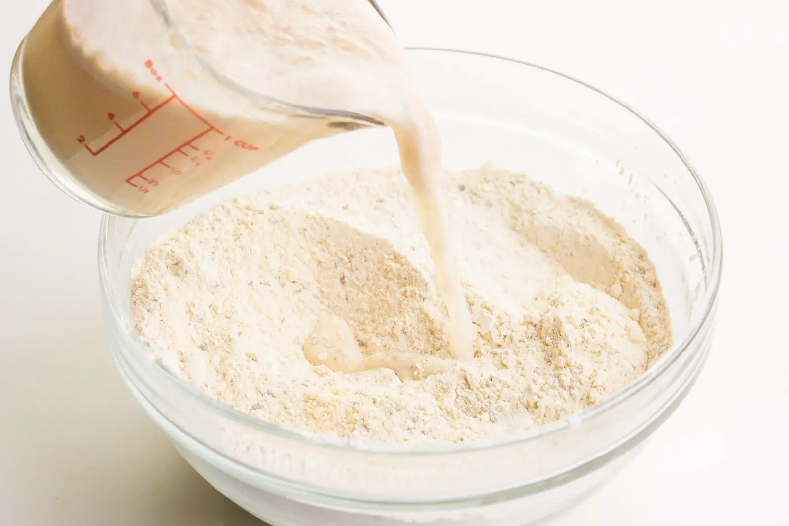 A milky yeast mixture is being poured into a bowl with a cassava flour mixture.