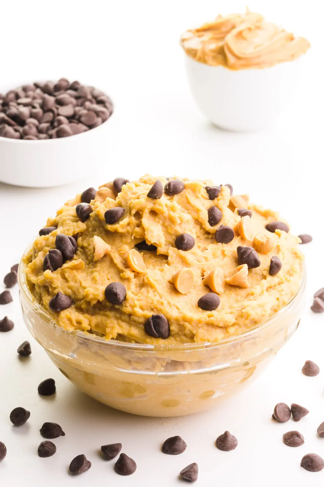 A bowl of cookie dough has chocolate chips around it. There's a bowl of chocolate chips and peanut butter behind it.