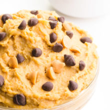 A bowl of chickpea cookie dough has chocolate chips on top. There is a bowl of chocolate chips behind it.