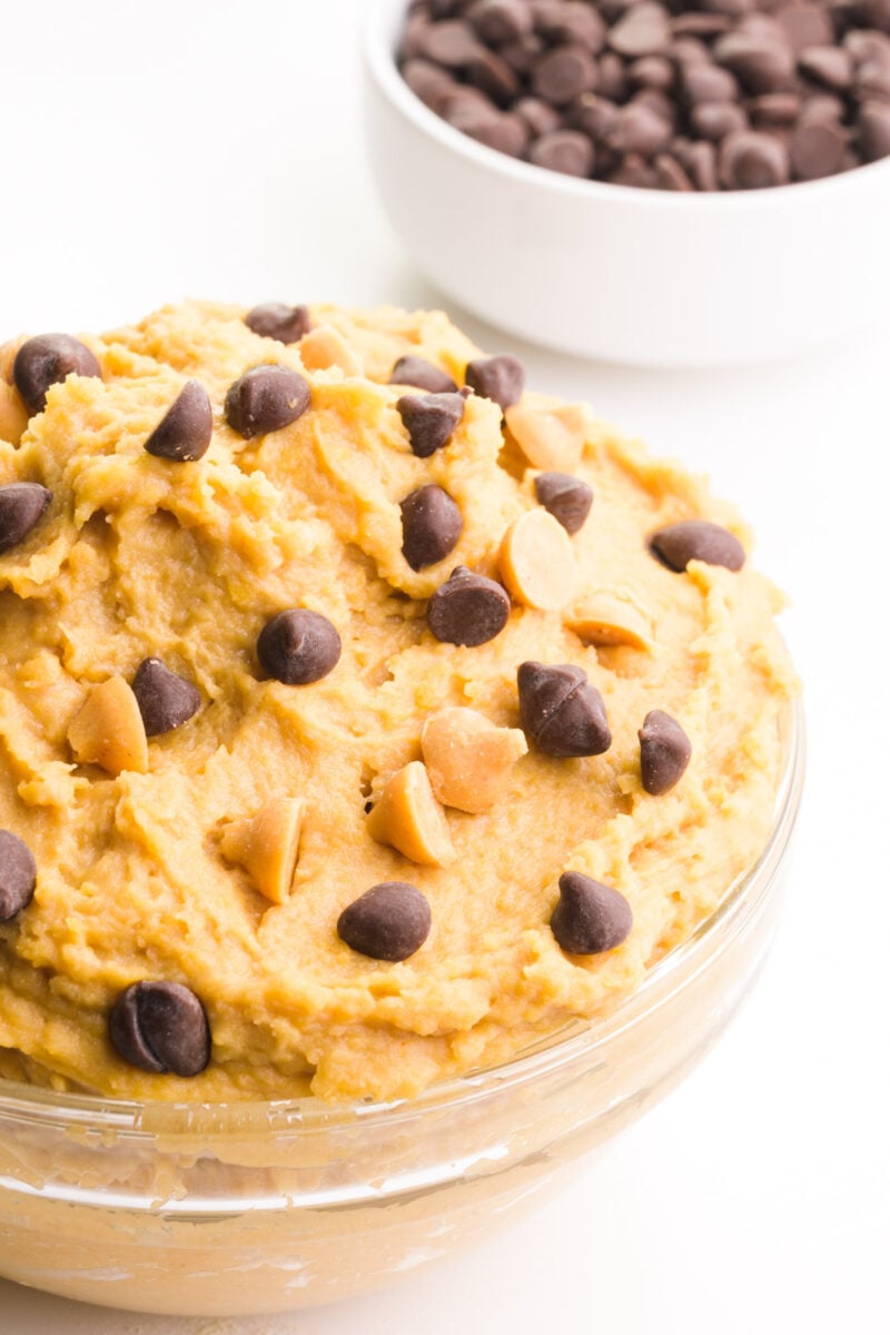 A bowl of chickpea cookie dough has chocolate chips on top. There is a bowl of chocolate chips behind it.