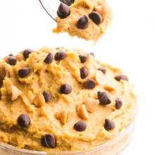 A hand holds a spoonful of cookie dough hovering over a bowl full of more cookie dough.