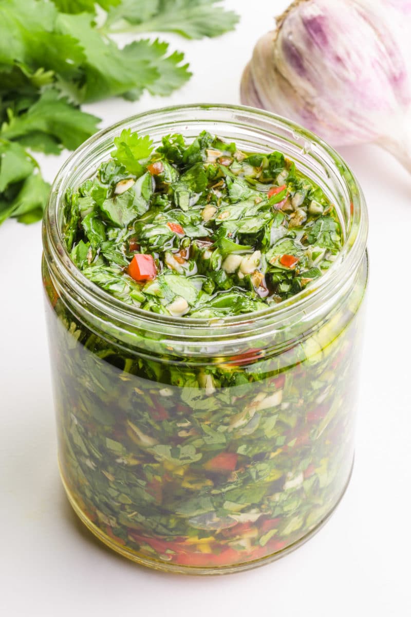 A glass jar holds chimichurri sauce. There is garlic and cilantro leaves in the background.