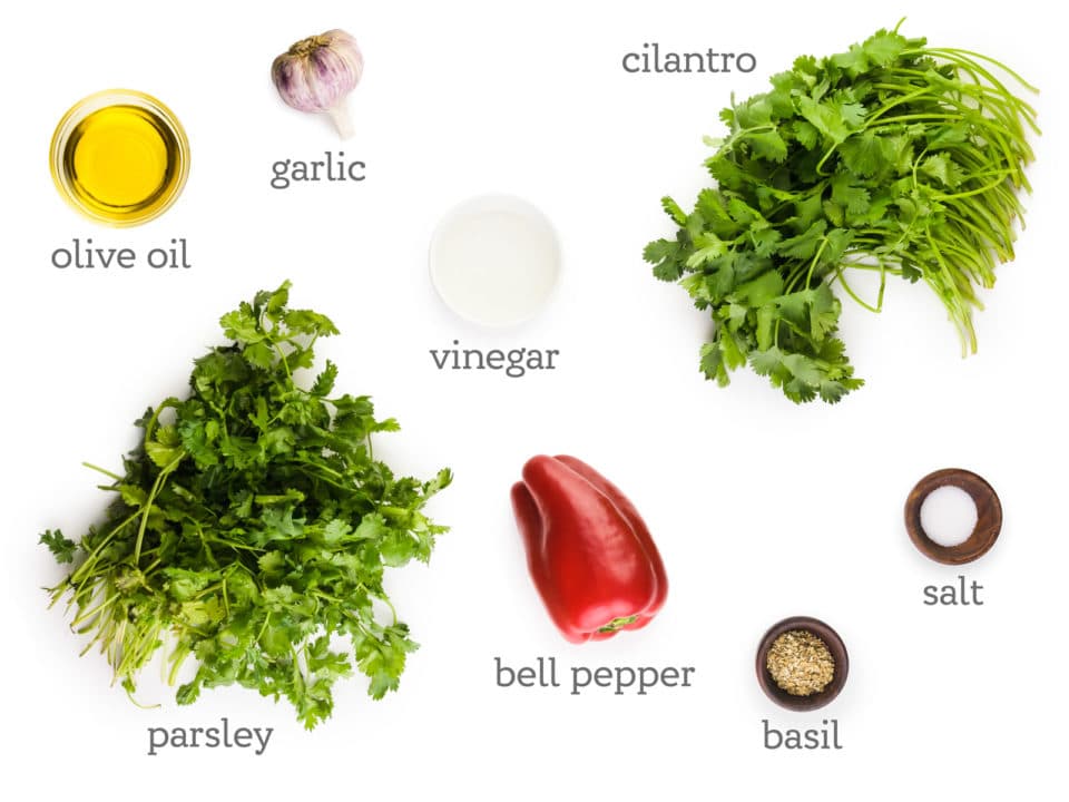 Ingredients are on a white table. The labels next to them read, cilantro, salt, basil, bell pepper, parsley, vinegar, olive oil, and garlic.