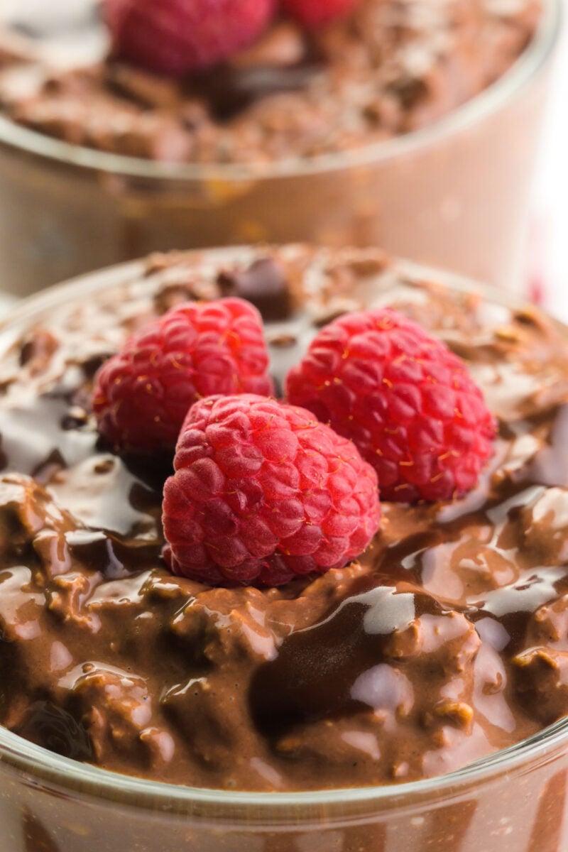 A bowl of chocolate overnight oats has raspberries on top. There's another bowl of oats the background.