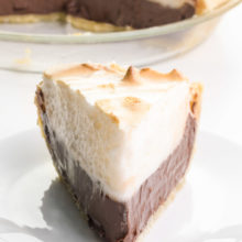A slice of chocolate pie sits on a plate. It's piled high with meringue. The rest of the pie is in a glass pie pan behind it.