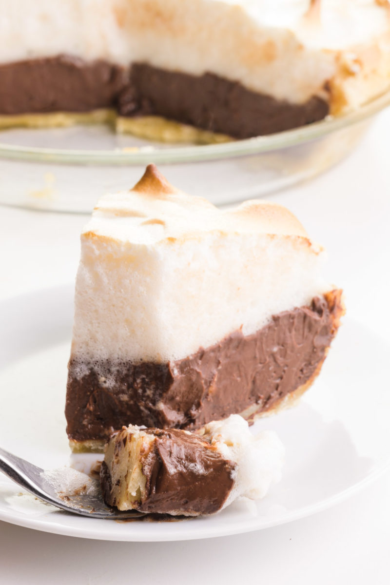 A slice of chocolate meringue pie has a bite taken out, sitting on a fork in front of the slice. The rest of the pie is behind it.