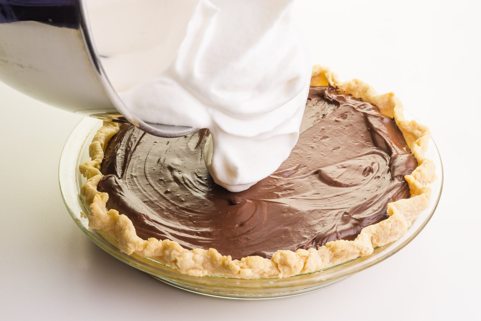 Vegan meringue is being poured from a mixing bowl onto a chocolate pie.