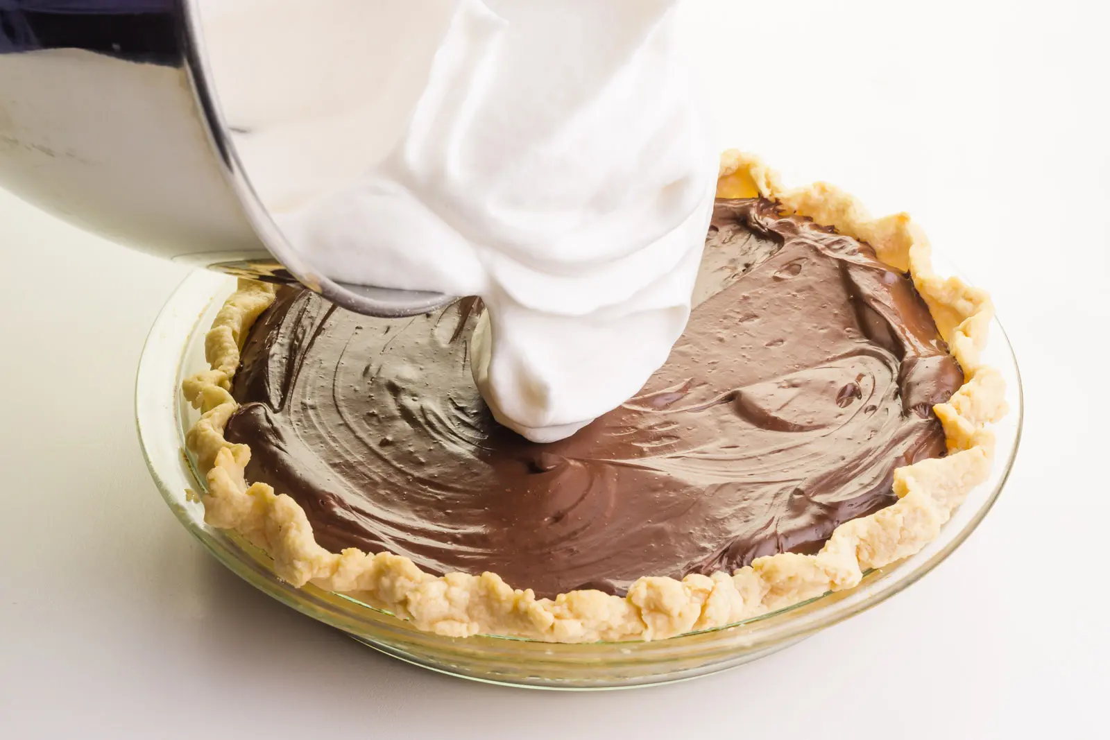 Vegan meringue is being poured from a mixing bowl onto a chocolate pie.