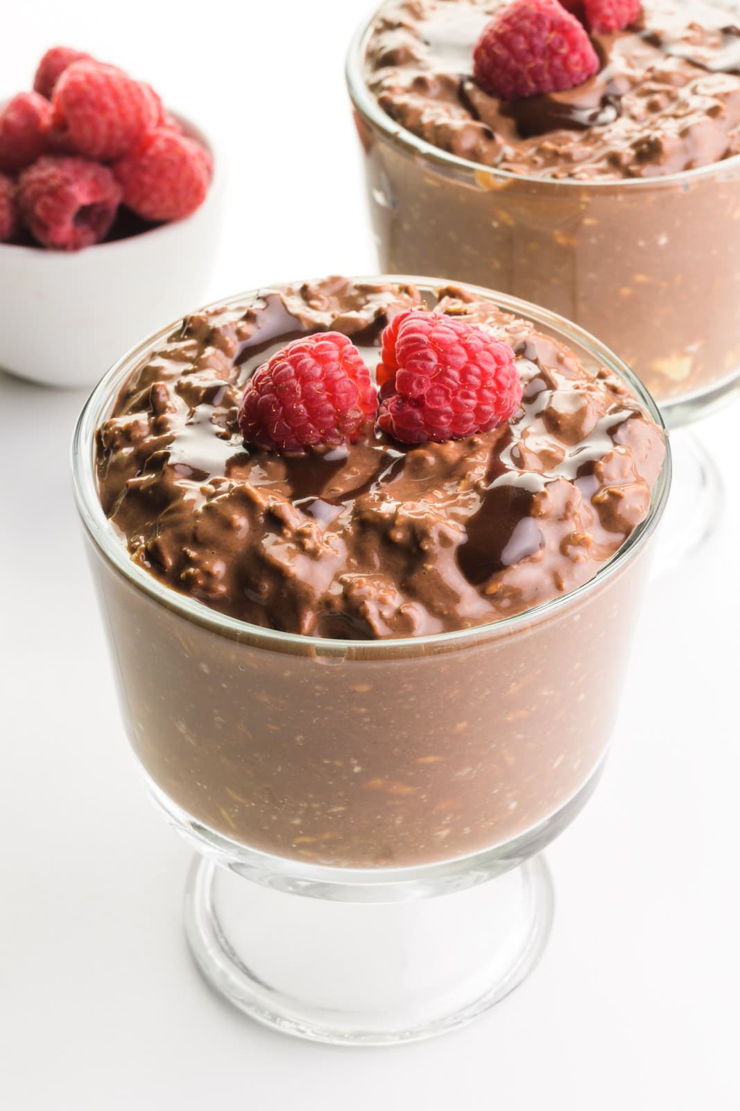 Two bowls of chocolate overnight oats have chocolate syrup and raspberries on top. There is a bowl of raspberries in the background.