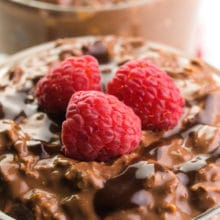 A bowl of chocolate overnight oats has raspberries on top. There's another bowl of oats the background.