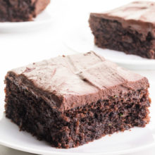 Three slices of vegan chocolate zucchini cake are on plates. They all are topped with chocolate frosting.