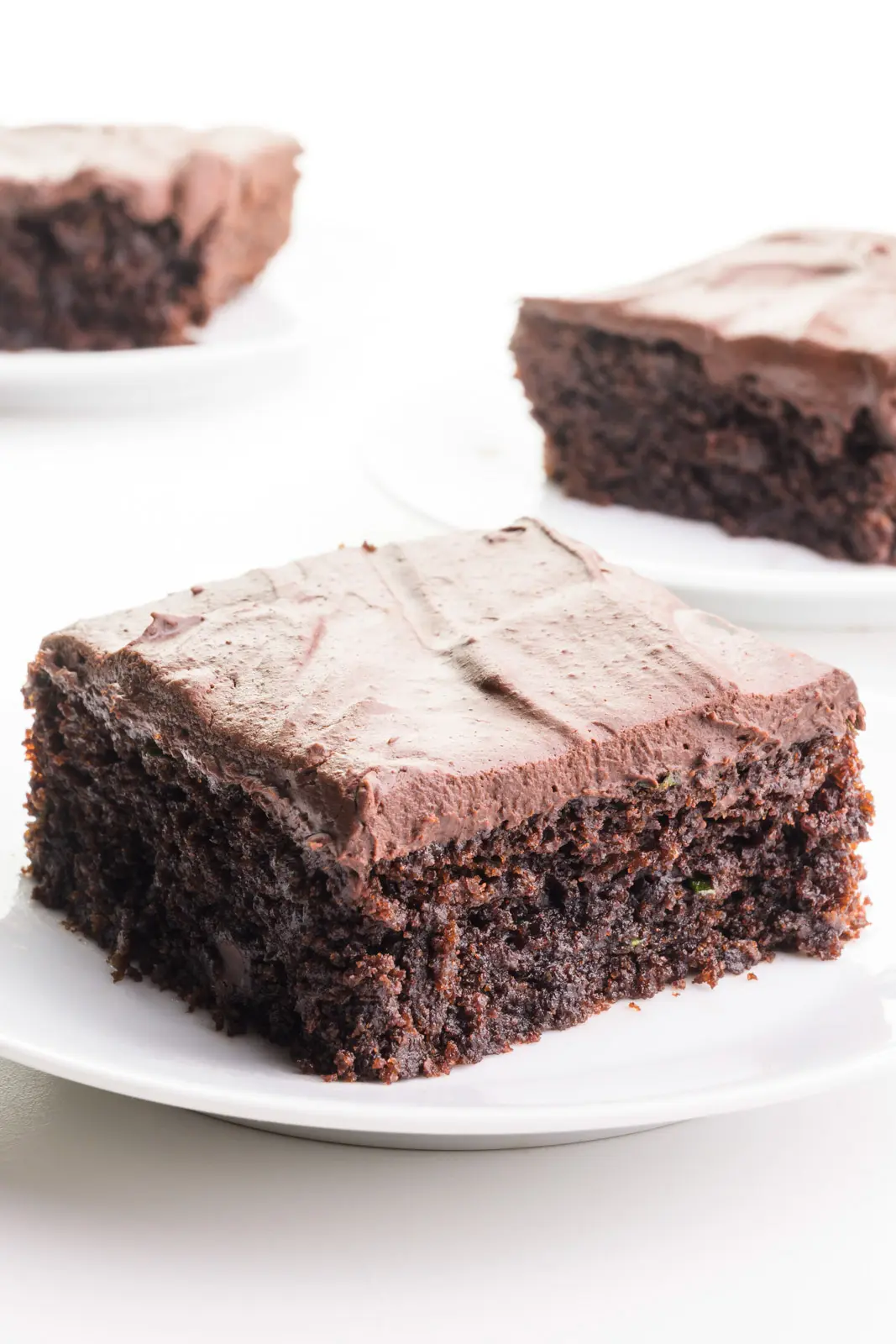 Three slices of vegan chocolate zucchini cake are on plates. They all are topped with chocolate frosting.