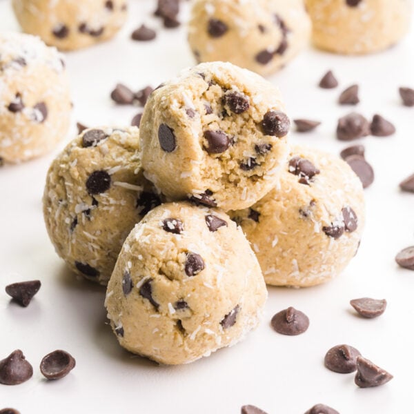 A stack of coconut protein balls shows the top one with a bite taken out. It's surrounded by chocolate chips and more protein balls in the background.