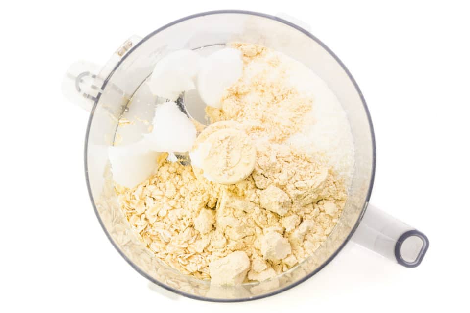 Ingredients are in the bottom of a food processor bowl, including protein powder, oats, and coconut oil.