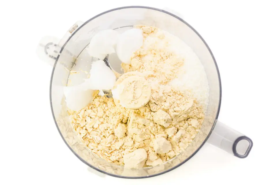 Ingredients are in the bottom of a food processor bowl, including protein powder, oats, and coconut oil.