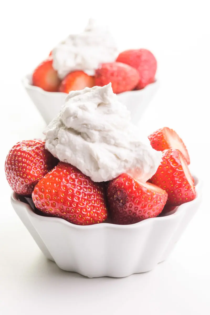 Two bowls of strawberries have coconut milk whipped cream on top. One is sitting in from the other.