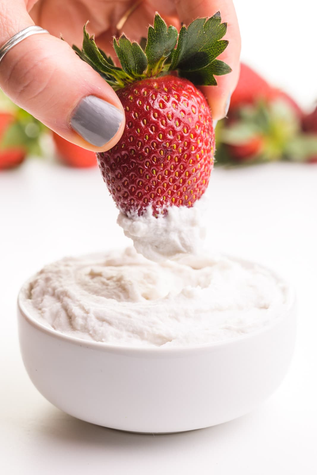 A hand holds a strawberry and is dipping it in a bowl of whipped coconut milk. There are more fresh strawberries in the background.
