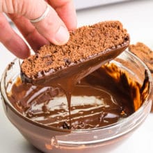 A hand holds a crisp chocolate cookie, dipping it in melted chocolate.