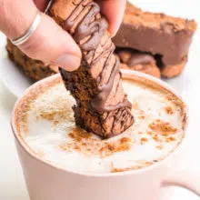 A hand is dipping a chocolate biscotti into a cup of hot chai tea.