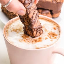 A hand holds a chocolate biscotti cookie and is dipping it in a pink mug with chai latte in it.