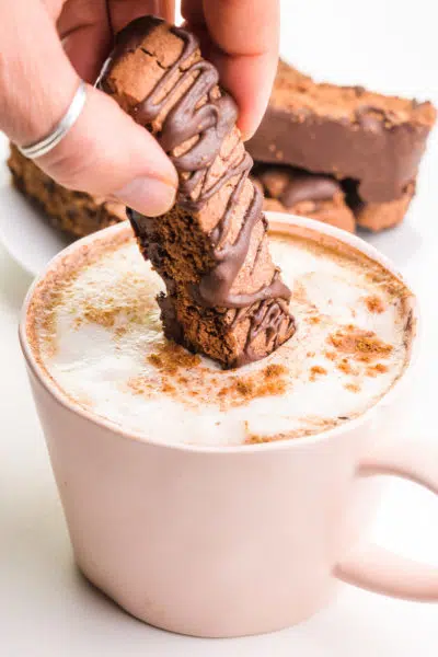A hand holds a chocolate biscotti cookie and is dipping it in a pink mug with chai latte in it.