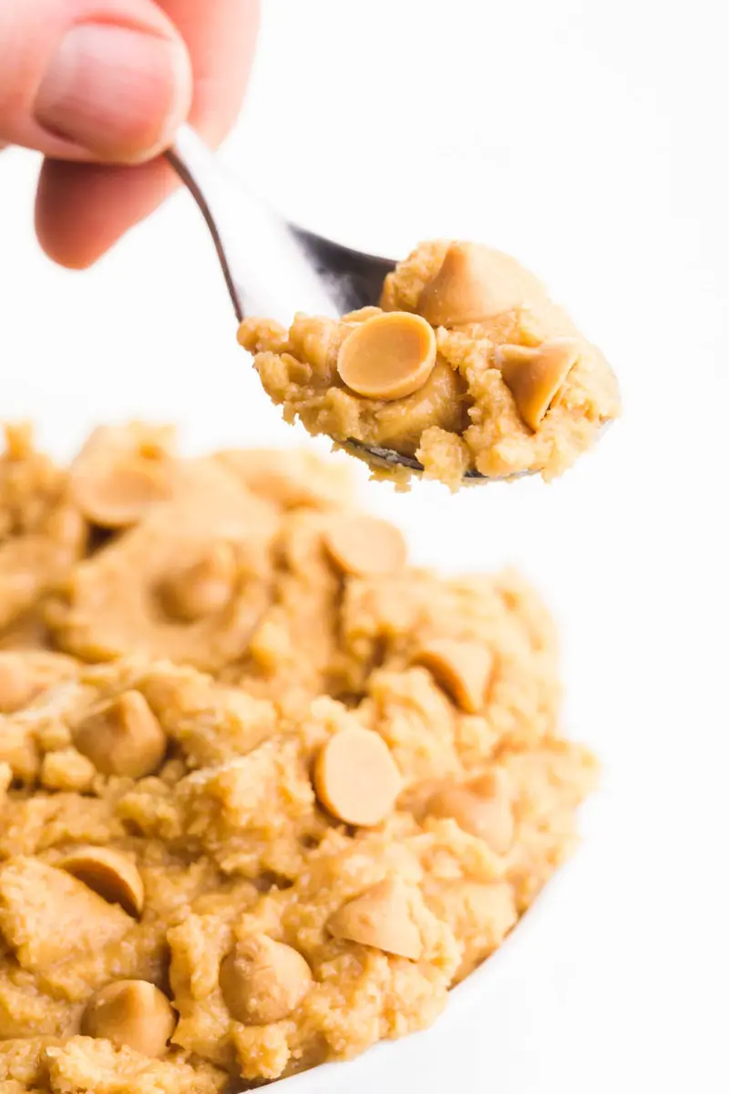 A hand holds a spoonful of edible peanut butter cookie dough in front of a bowl with more of it.