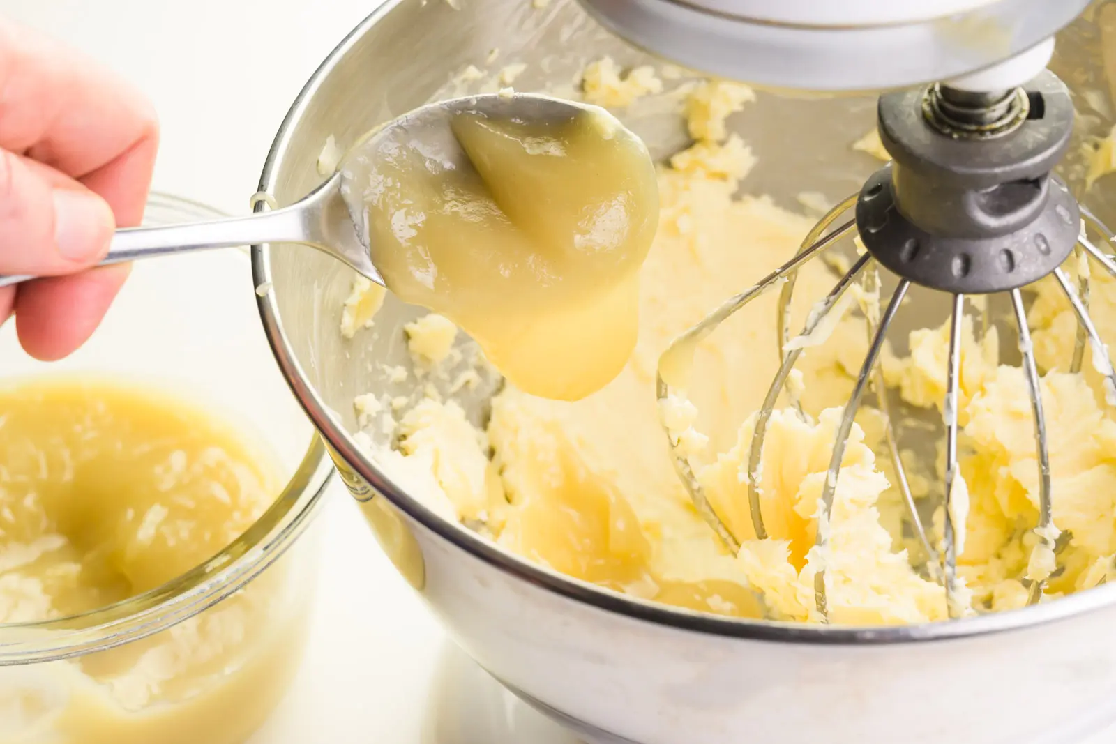 A hand holds a spoon dripping a creamy mixture in with whipped butter. A bowl of the creamy mixture sits beside the mixing bowl.