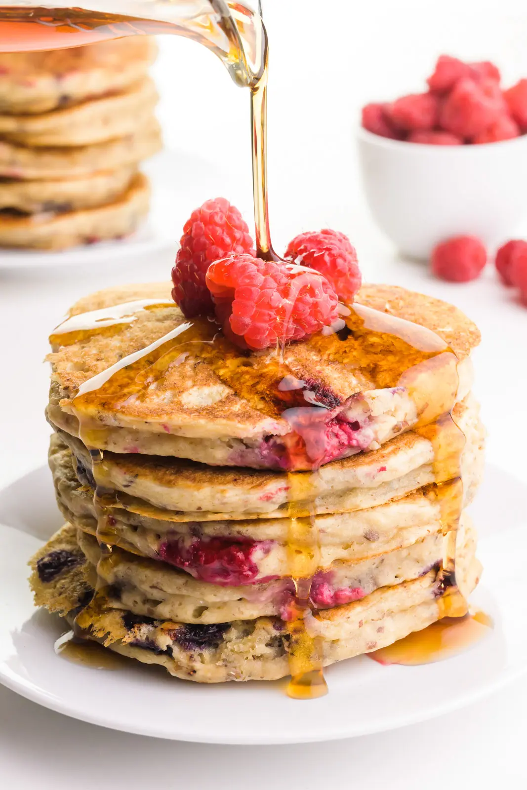 Syrup is being poured over fruity pancakes with raspberries on top. There's another stack in the background and a bowl full of raspberries.