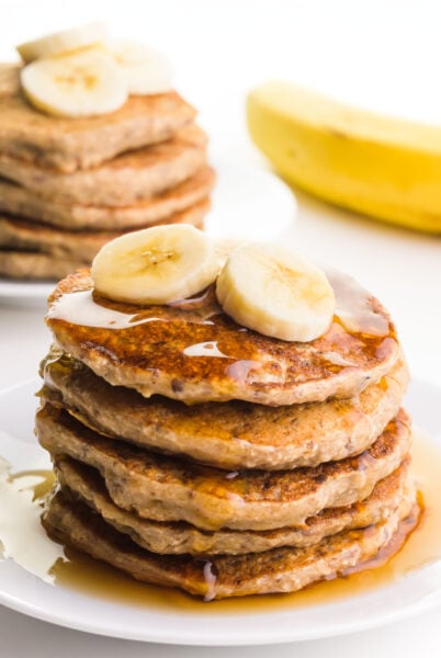 A plate holds several gluten-free banana pancakes with syrup on top. There are sliced bananas on top of the stack. There's another stack of pancakes on a plate next to a banana in the background.