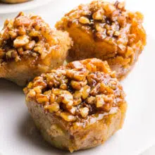 Three gluten-free sticky buns are on a plate. There's another plate of these rolls barely visible in the background.